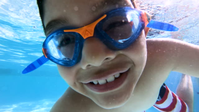 High quality stock video of a young 7-year-old boy making hand signs underwater looking into the camera in a bright, clean swimming pool.