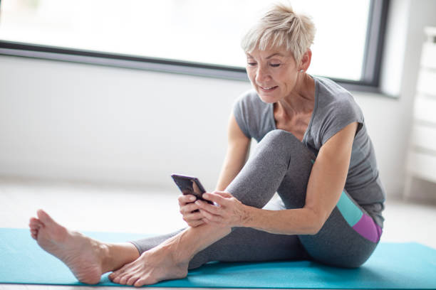 istock Modern short blonde hair mature woman using mobile phone while relax after exercise in corner of room with window at background 1128432746