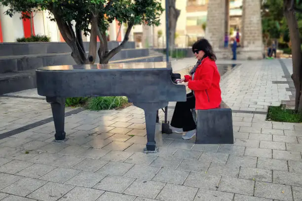 A woman pianist in a red jacket plays the grand piano in the street. The piano is a weatherproof instrument mounted in the street and available to any passer-by.