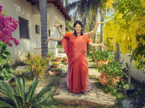 Two Caucasian dancers depict Lord of the Dance or Nataraja forming their hands in a symbol of the fertility. They stand on the wide stone step among flowers and palm trees. The frontwoman is in an orange sari.