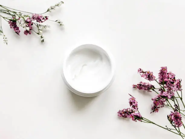 Skincare product in a white jar is standing on a white backdrop decorated with small flowers