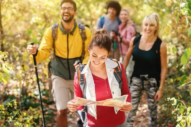 Photo of Smiling beautiful brunette holding map and leading the rest of hikers through woods in autumn.