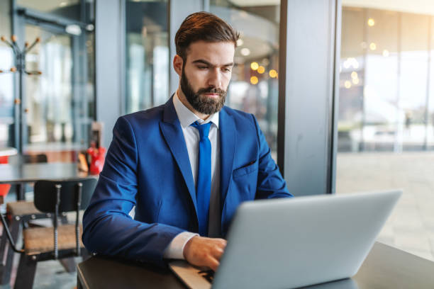 istock Bearded businessman in formal wear with serious facial expression using laptop while sitting in cafeteria next to window. 1128536214