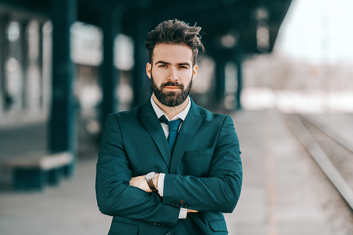 Portrait of serious Caucasian bearded businessman in formal wear standing at train station with arms crossed.