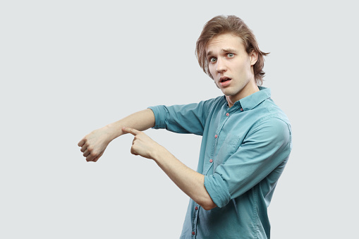 Portrait of worry handsome long haired blonde young man in blue casual shirt standing and showing time out gesture and looking looking at camera. indoor studio shot, isolated on light grey background.