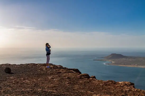 Photo of A woman stands on the edge of a cliff and looks at a nearby island in the ocean.