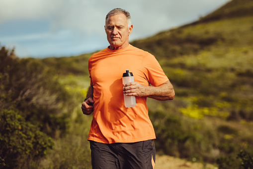 Fitness man jogging outdoors holding a water bottle. Portrait of a senior man running outdoors with a hill in the background.