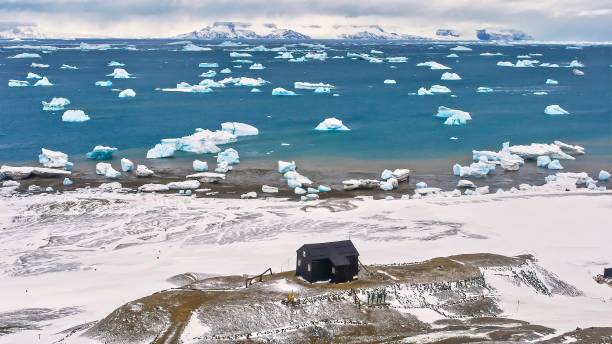 High angle landscape view taken on Snow Hill Island, Antarctica, showing a small black hut on a hill surrounded by ice and snow, and Admiralty Sound dotted with icebergs, and James Ross Island in the distance. A dramatic view of an Antarctic landscape, with a small black building looking small against the snowy coastline of Snow Hill Island, and the sea dotted with many floating icebergs. In the background another island is visible. houses in antarctica stock pictures, royalty-free photos & images