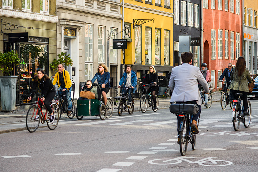COPENHAGEN; DENMARK - MAY 03; 2018: Group of people riding bicycles in old city center. A man sitting in front cart of wheelbarrow cargo bike. Movement in both directions, road markings cycle lane. Street style