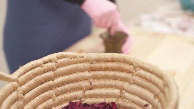 Close-up side view of a basket, full of dried flowers. The camera moves and shows how the girl grinds dried rose petals into powder in a copper mortar with pestle.