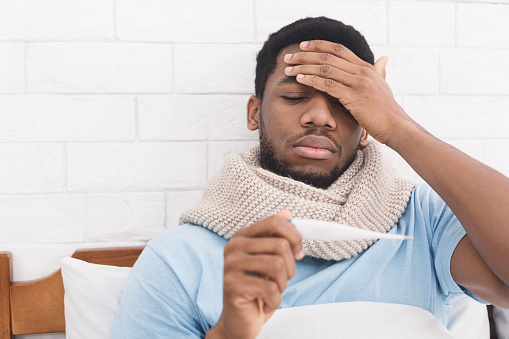 Sick african-american man having fever, measuring body temperature with thermometer, touching forehead in bed, copy space