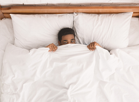 African-american man in bed hiding under blanket, top view, copy space