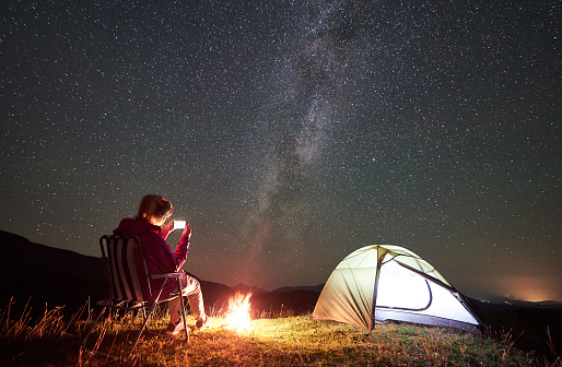 Female hiker having a rest at summer night camping in the mountains beside campfire and glowing tourist tent. Young woman sitting on chair, taking picture of night sky full of stars and Milky way.