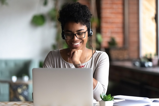 Black woman smart student girl sitting at table in university cafe alone wearing glasses looking at computer screen using headphones listening online lecture improve language skills having good mood