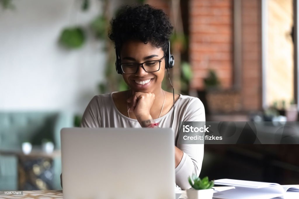 Smart student learning using internet and headphones Black woman smart student girl sitting at table in university cafe alone wearing glasses looking at computer screen using headphones listening online lecture improve language skills having good mood Internet Stock Photo