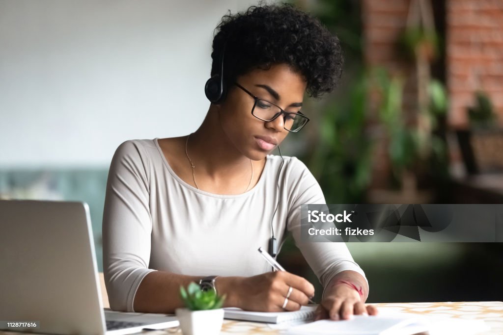 Serious african woman studying using internet holding pen writing notes Serious african female smart student sitting at table wearing headphones listening online lecture writing notes on textbook. Self education and knowledge improvement using modern technologies concept Efficiency Stock Photo