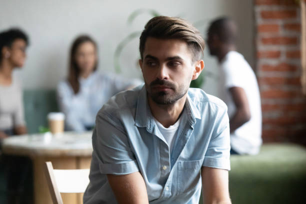Frustrated lonely shy guy sitting apart of others teenagers Different friends sitting in cafe after study spending time together, focus on frustrated lonely shy guy sitting apart, separately of others teenagers feels unhappy having problems with communication indifference stock pictures, royalty-free photos & images