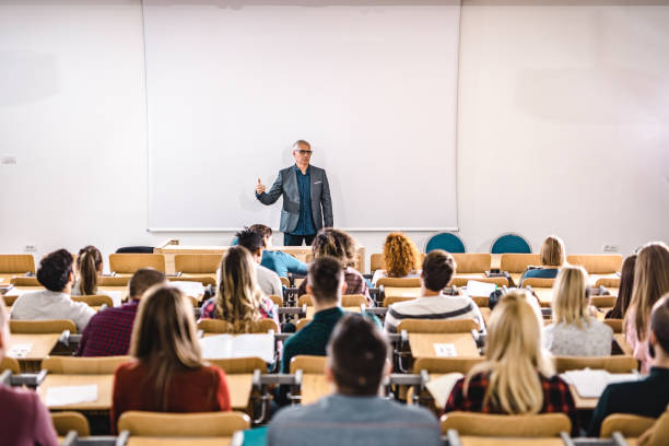 Senior teacher talking to large group of college students in amphitheater. Mature professor giving a lecture in front of projection screen at lecture hall. Copy space. classrooms stock pictures, royalty-free photos & images