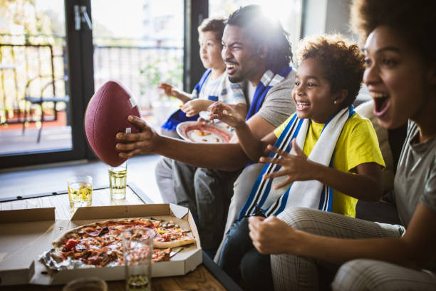 Cheerful black family cheering while watching rugby match on TV at home. Young joyful black family having fun while cheering for their favorite American football team at home. sports event stock pictures, royalty-free photos & images