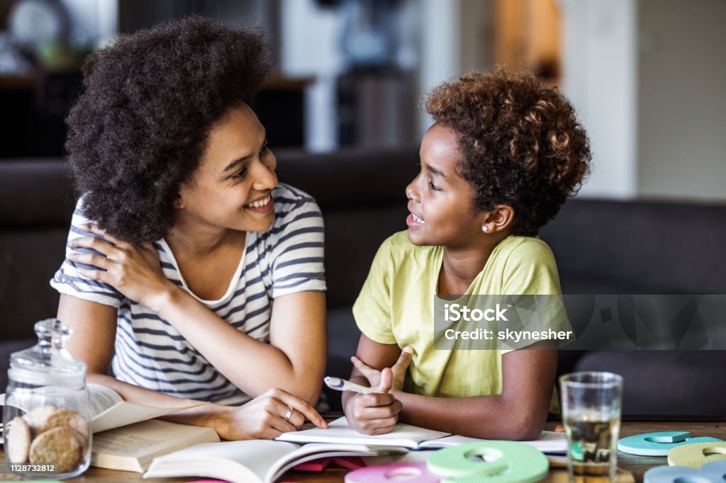 Happy African American mother assisting her daughter with homework. Small African American girl talking to her mother while writing her homework in the living room. Child Stock Photo