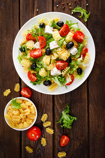 Italian pasta salad with fresh tomato, cheese, lettuce and olives on wooden background. Mediterranean cuisine. Cooking lunch.  Healthy diet food