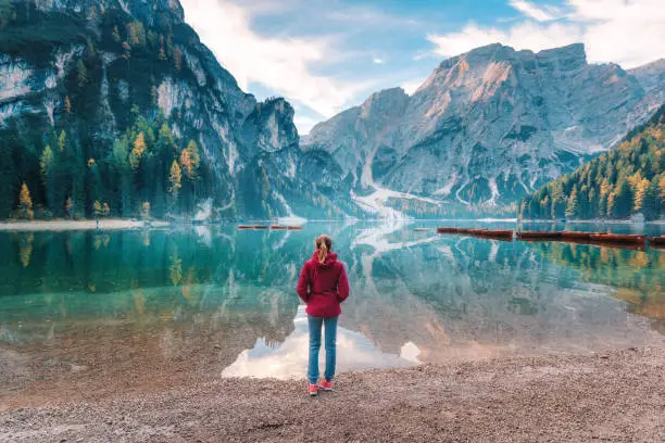 Woman in red jacket is standing on the coast of Braies lake at sunrise in autumn. Dolomites, Italy. Landscape with girl, famous lake with beautiful reflection in water, trees, sky with clouds. Travel