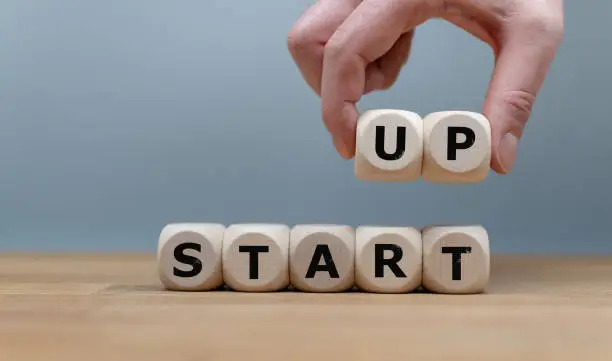 Cubes form the words "START UP" while to fingers lift the letters "UP" in the air. Cubes are on a wooden table in-front a grey background.