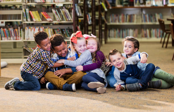 Children in library, girl with down syndrome, group hug A group of six multi-ethnic children sitting together on the floor of a school library, hugging. The girl sitting 3rd from the right, has down syndrome. down syndrome photos stock pictures, royalty-free photos & images