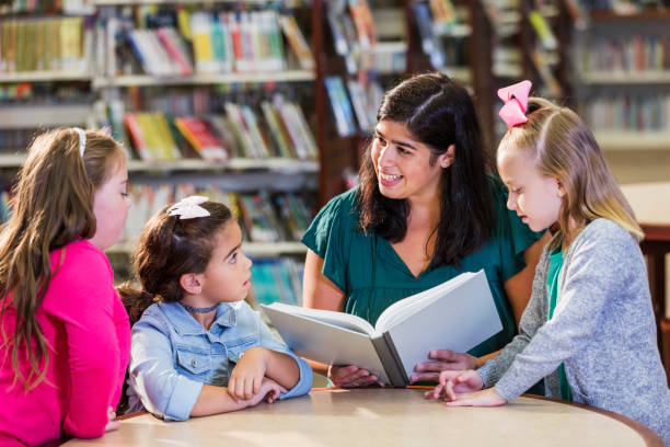 istock Hispanic teacher and three girls in library reading book 1128757590