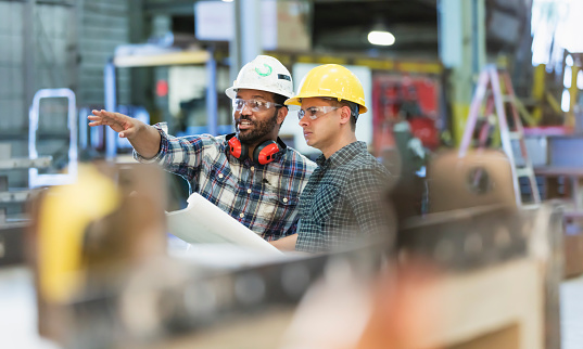 Two multi-ethnic workers in their 30s talking in a metal fabrication plant wearing hardhats and protective eyewear. The man pointing is African-American and his coworker is Hispanic.