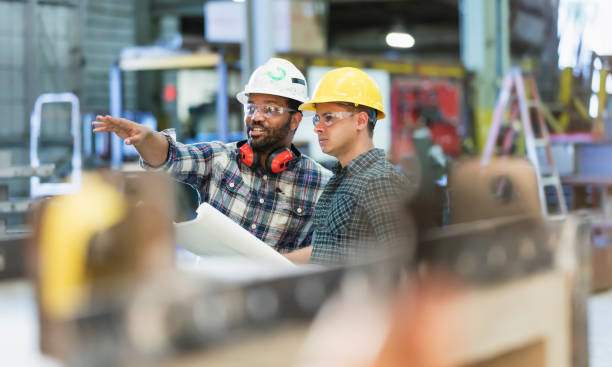Multi-ethnic workers talking in metal fabrication plant Two multi-ethnic workers in their 30s talking in a metal fabrication plant wearing hardhats and protective eyewear. The man pointing is African-American and his coworker is Hispanic. hard hat stock pictures, royalty-free photos & images