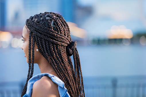 A young mixed race woman in her 20s in the city, standing outdoors on the waterfront. She has beautiful long black hair, cornrow braids tied back with a hair band.