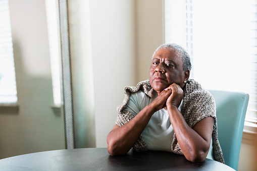 A lonely senior African-American woman in her 70s sitting at home at a table by a window. She is looking at the camera with a serious expression, elbows on the table and hands clasped.