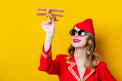 Portrait of charming vintage stewardess wearing in red uniform and sunglasses with airplane. Isolated on yellow background.