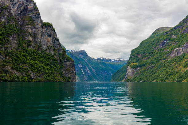 The mountains in the Geirangerfjord are captured on a cloudy summer day. The slopes of the mountains are covered with the green vegetation, and the landscape is reflected in the water.