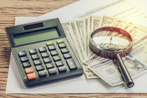 Tax, search for return or financial report review concept, magnifying glass on pile of US Dollar banknotes money with calculator on graph and chart profit and loss report on wood table.