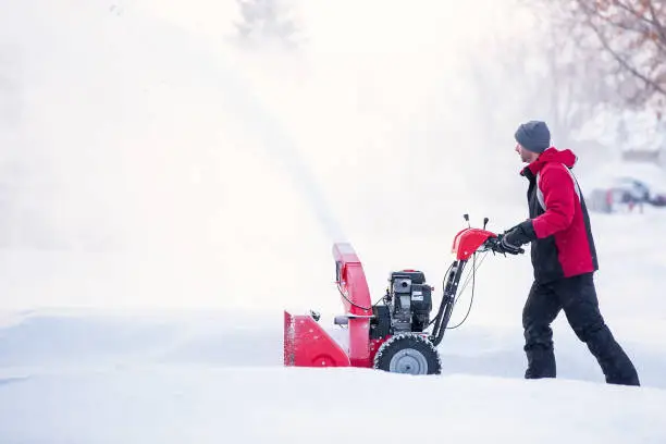 Side view of a man using a snow blower to remove snow from a driveway on a winter day.