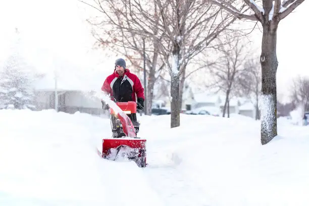 Front view of a man using a snow blower to remove snow from a sidewalk in town on a winter day.