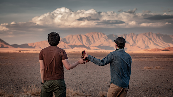 Two Asian man traveler clinking bootle drinking beer while looking at scenery during the sunset in Namibia, Africa. Friendship, travel and celebration concepts