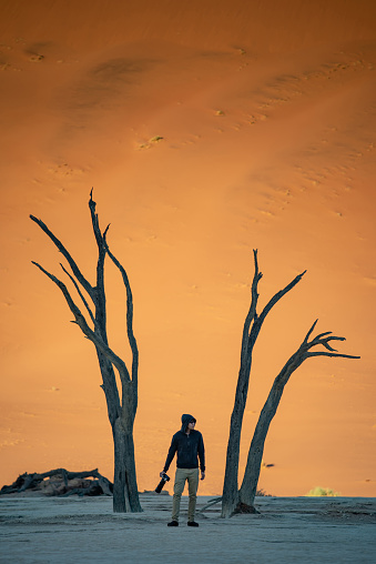 Young Asian man traveler standing between dead trees in deadvlei (Sossusvlei) during sunrise, famous natural landmark in Namib desert of Namibia, Africa