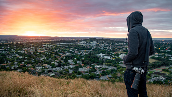 Hoodie man photographer and traveler watching sunrise over Auckland city at Mount Eden view point in Auckland, North Island, New Zealand. Travel Oceania. Success or achievement concepts