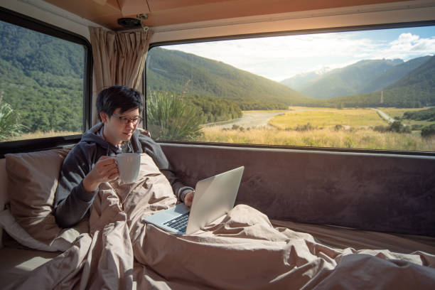 istock Young Asian man traveler drinking hot coffee while working with laptop computer on the bed in camper van with mountain scenic view through the window. Digital nomad or freelance on road trip concept 1128825797