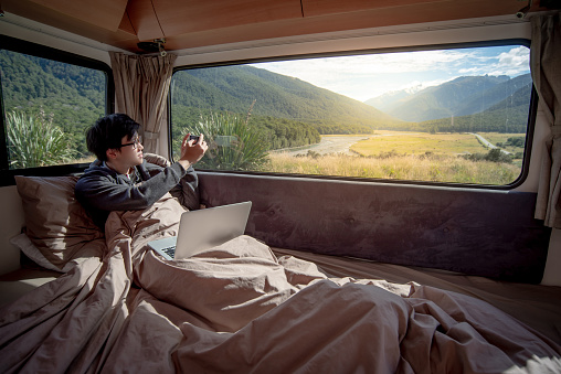 Asian man traveler enjoy taking photo of mountain scenery through the window by smartphone while staying in the blanket in camper van in the morning. Road trip in summer of South Island, New Zealand.