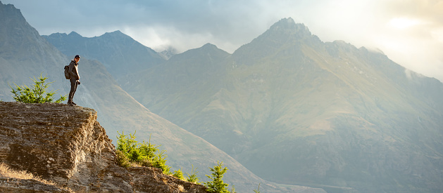 Asian man traveler and photographer standing on the rock among mountain landscape during golden hour sunset in Queenstown, South Island, New Zealand. Travel Oceania. Success or achievement concept