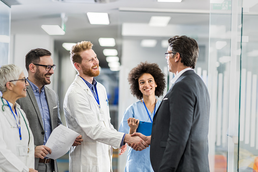 Happy doctor shaking hands with a businessman while standing with their colleagues in the hospital.
