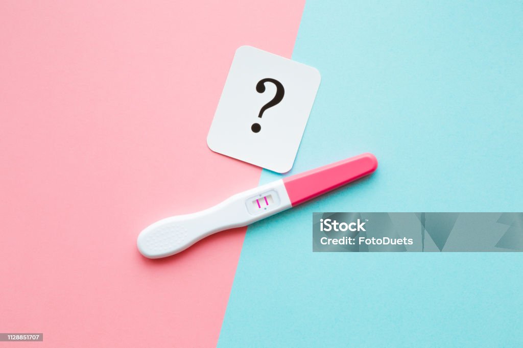 Pregnancy test with two stripes and white card of black question mark. Pastel blue and pink background. Positive result. Guessing unborn baby gender. Closeup. Pregnancy Test Stock Photo