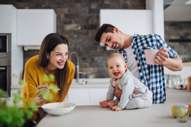 A young family at home, a man holding a baby and a woman feeding her. A portrait of young family standing in a kitchen at home, a man holding a baby and a woman feeding her with a spoon. family at home stock pictures, royalty-free photos & images