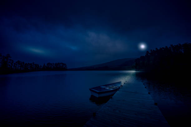 Night scene of a Moored rowboat on a lake next to a wooden jetty overlooking mountains and woodlands with the moon behind the clouds reflecting on the water in the Cape Winelands South Africa