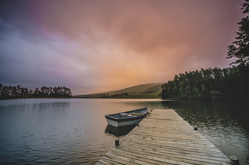 A Moored rowboat on a lake next to a wooden jetty overlooking mountains and woodlands on a cloudy morning with orange coloured sunrise at dawn Cape Winelands South Africa