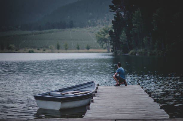 man kneeling looking out on a wooden jetty on a beautiful lake with a moored rowboat in nature - loch rowboat lake landscape imagens e fotografias de stock